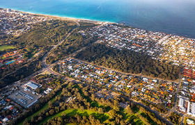 Aerial coastal view of a suburban area bordered by the ocean, showing a curved sandy shoreline with blue water on one side and dense residential housing on the other.