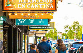 A street scene showing a small shopping strip with several storefront signs, including a large orange sign reading “Hermanos en Cantina” above a shop entrance.