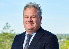 Head-and-shoulders portrait of a person wearing a dark blue suit jacket, light-colored shirt, and patterned tie, standing outdoors with a blurred background of greenery and sky. 