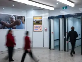 Indoor airport arrivals area with white walls and tiled flooring, featuring glass exit doors marked “Way Out.” A digital advertising screen on the wall displays a “Do more you at Scarborough Beach”