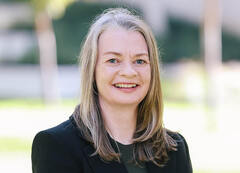 Head-and-shoulders portrait of a person with long, straight, light-colored hair wearing a black blazer over a dark top, standing outdoors.