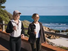 Two women walking along the coast with coffee