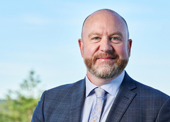 Head-and-shoulders portrait of a person wearing a blue check-pattern suit jacket, light-colored shirt, and patterned tie, standing outdoors. 
