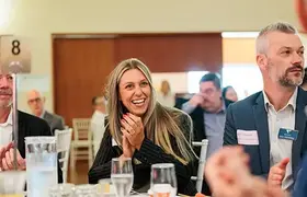 Business event attendees seated at round tables listening to a speaker, with one person in the foreground clapping and glasses and table settings visible