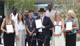 Group of eight people outdoors holding framed certificates, several wearing medals. One person in front stands with a walking frame. Trees and a building are visible in the background.