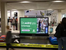 Airport baggage claim area with a luggage carousel in the foreground and travellers standing nearby. A large digital display on the carousel shows a green “Do more you in Karrinyup” 