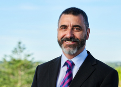 Head-and-shoulders portrait of a person with short dark hair and a neatly trimmed beard, wearing a black suit jacket, light blue shirt, and a pink-and-purple striped tie.