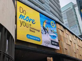 Large outdoor digital billboard mounted on a modern city building displays a bright yellow and blue “Do more you at Scarborough Beach” advertisement with an image of people surfing on ocean waves.