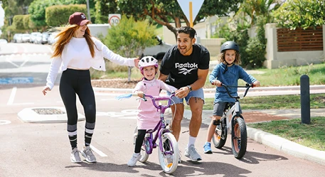 Two children riding bicycles while two adults walk beside them on a tree-lined suburban street, promoting active and healthy lifestyles.