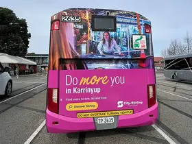 Rear view of a bus stopped in a transport area, covered in a bright pink advertisement reading “Do more you in Karrinyup” with smaller text “Find more to see, do and love” and “Discover Stirling,”
