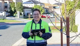 Person in a high-visibility jacket holding a tablet while standing on a suburban street footpath.