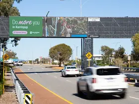 Cars drive along a multi-lane road beneath an overhead pedestrian bridge in a suburban area. The bridge features a large green advertisement reading “Do more you in Karrinyup” alongside smaller text “