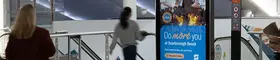 An indoor public walkway with tiled floors and glass railings features a freestanding digital advertising display. 