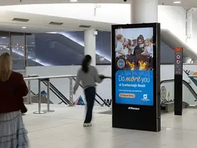 An indoor public walkway with tiled floors and glass railings features a freestanding digital advertising display. 