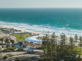 Panoramic view of a coastal beach with turquoise ocean waves rolling toward a sandy shoreline, bordered by a park with green lawns and tall pine trees.