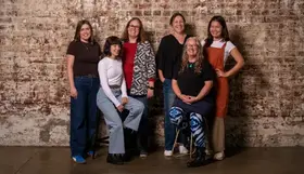 Six artists pose in a studio against a weathered brick wall, with one seated on a stool in front and others standing close behind in casual clothing.